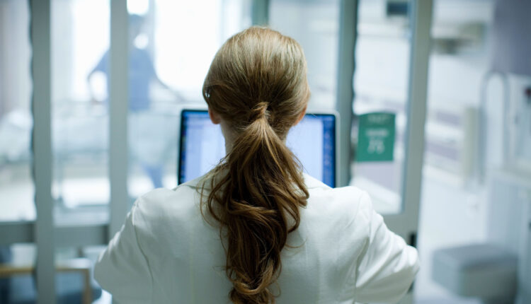 A photo of a woman seen from behind working in a medical setting on a laptop.