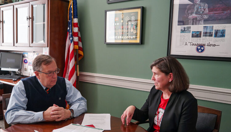 Barb Dentz (right), sits beside state representative, Sam Whitson (left), in an office room. The walls are an olive green and are adorned by framed medals. An American flag stands in the corner, behind them.