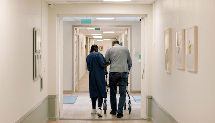A photo of a female nurse helping an elderly man using a walker down a corridor.