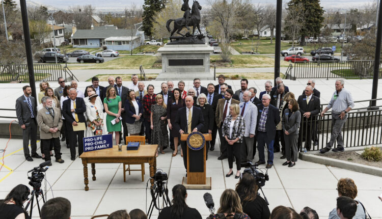 Gov. Greg Gianforte, surrounded by Republican lawmakers, speaks at a bill signing ceremony on the steps of the State Capitol, in Helena, MT. Press huddles in front of the podium he speaks from with microphones and cameras.