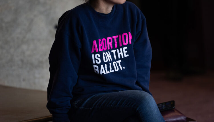 A woman is photographed from the chin down, with the focus on her sweatshirt. The shirt reads, "ABORTION IS ON THE BALLOT."