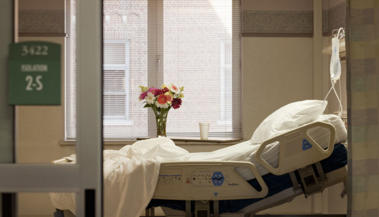 A photo showing a vacant hospital room. The bed is empty and there are flowers on the window sill.